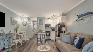 Et moderne køkken med hvide skabe, en mikrobølgeovn og et køleskab. Der er et spiseområde med et bord og stole, samt en stue med en beige sofa og dekorative puder.