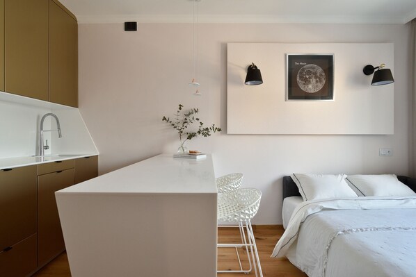 A modern, minimalist bedroom with a white bed, a small white table with chairs, and a wall-mounted artwork. - Warszawianka Studio (Warsaw)