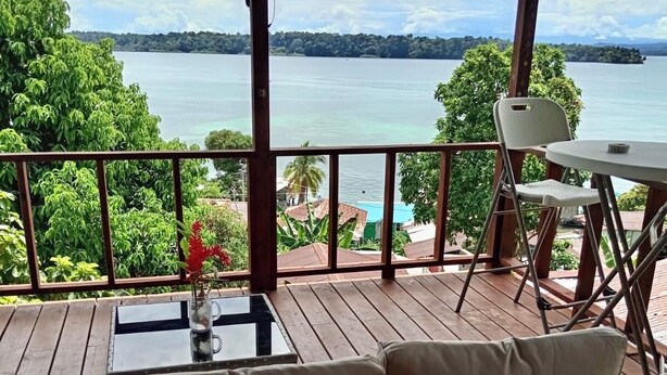 Un espacio al aire libre techado con mobiliario de mimbre, una mesa con un jarrón y vistas a un lago y tierras lejanas.