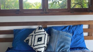 Una cama con ropa de cama azul y una almohada estampada, cabecera de madera y ventana con vista a follaje.
