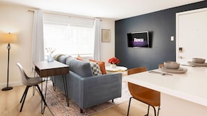 Ein modernes Wohnzimmer mit einer Couch, einem Esstisch und Stühlen, einem flachbildfernseher an der Wand und einem Fenster mit Vorhängen.