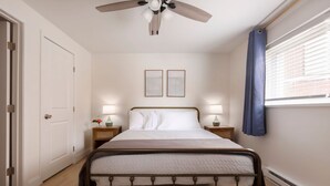 Ein Schlafzimmer mit Bett, Deckenventilator, Nachttischlampen und einem Fenster mit Vorhängen.