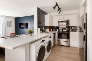 A modern kitchen with a built-in washer and dryer, stainless steel appliances, and a flat-screen TV mounted on the wall. - Homey Haven in Tower Grove Near Major Hospitals
(St. Louis)