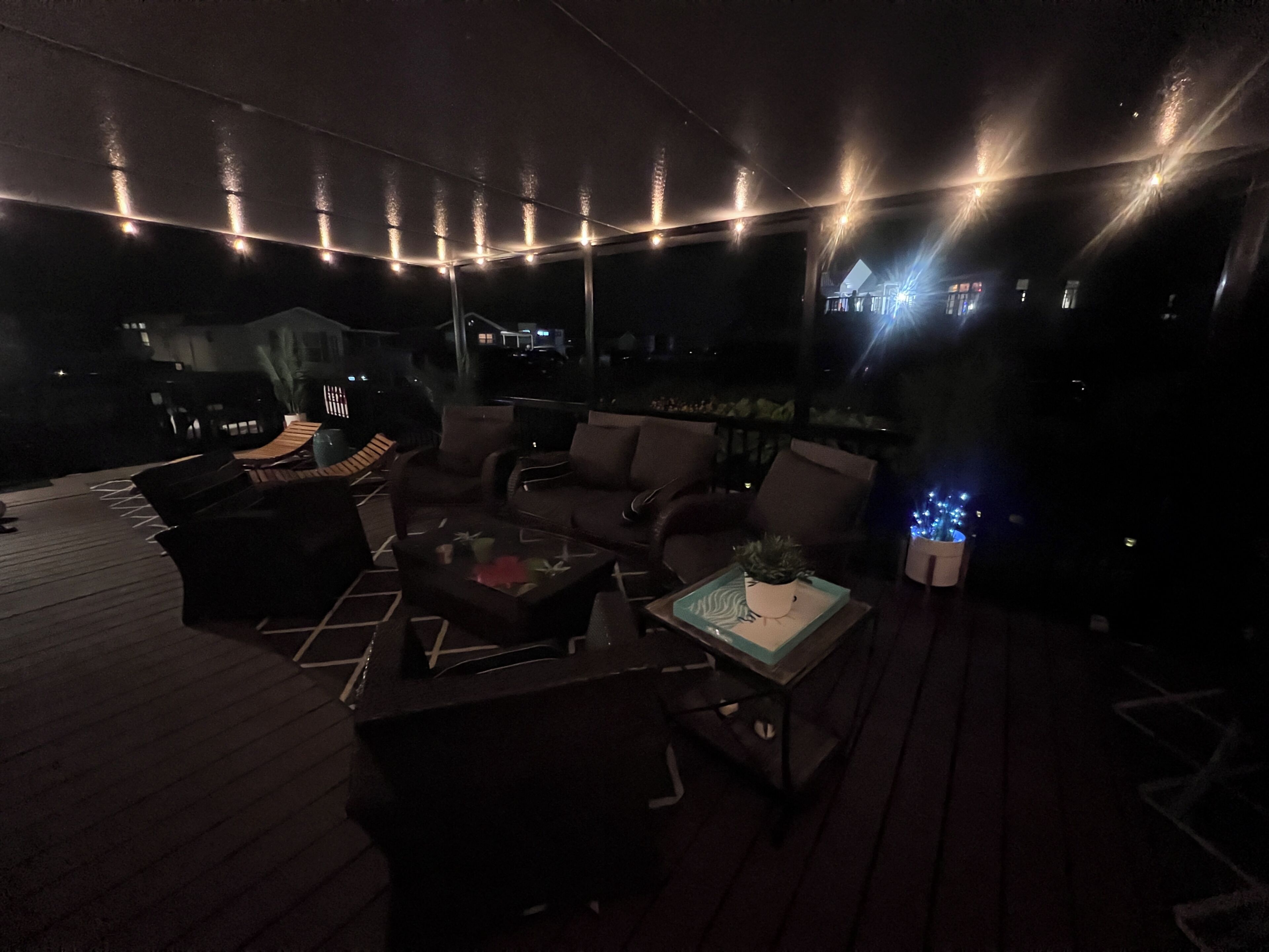 Un espace extérieur aménagé en terrasse, avec un sol en bois, des canapés et une table basse, et des guirlandes lumineuses suspendues au-dessus.