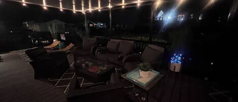 Un espace extérieur aménagé en terrasse, avec un sol en bois, des canapés et une table basse, et des guirlandes lumineuses suspendues au-dessus.