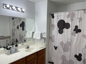 Une salle de bain avec un rideau de douche à l’effigie de Mickey Mouse, un meuble-lavabo avec un miroir et un sèche-cheveux fixé au mur.