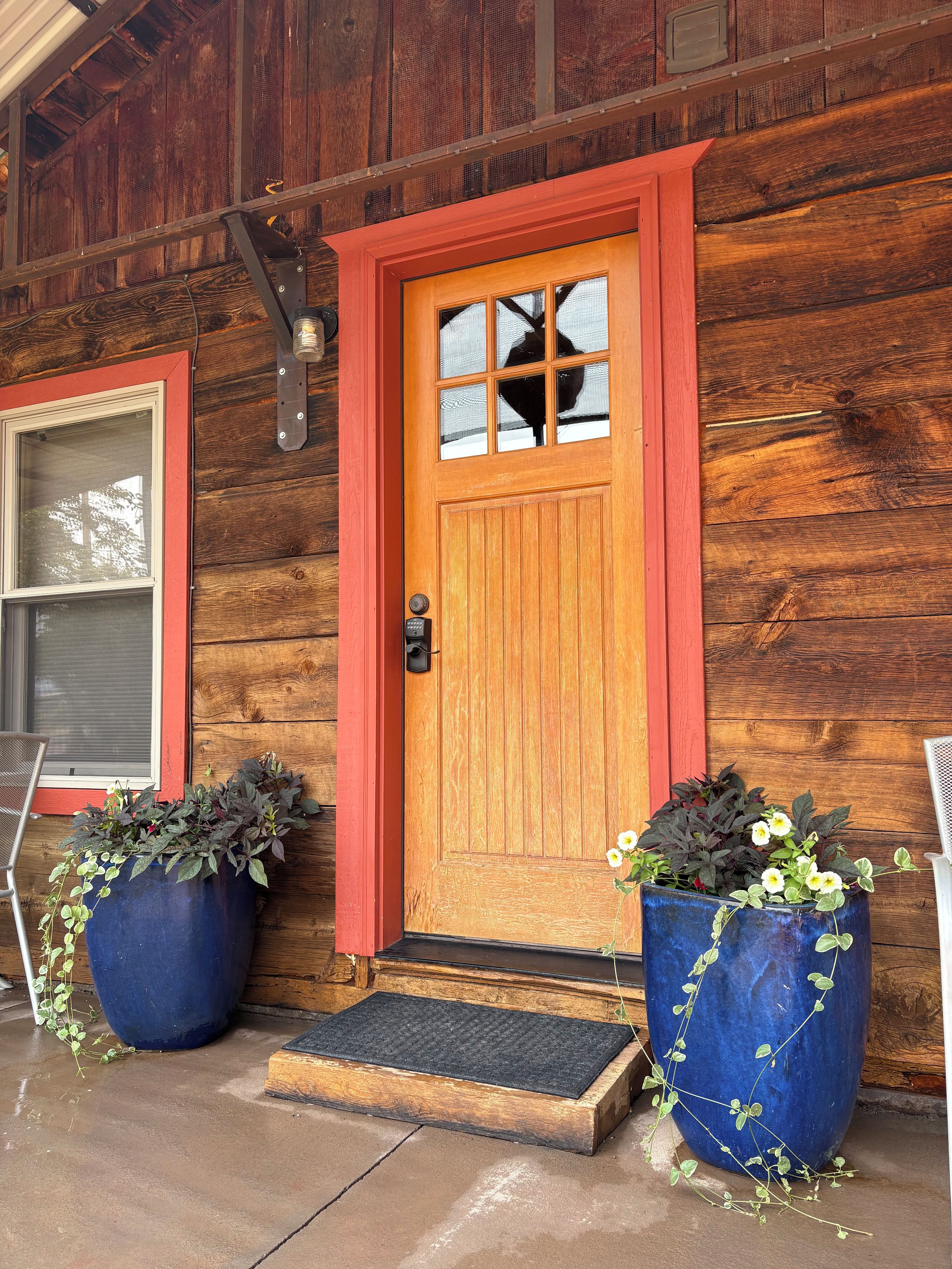 Une maison en bois avec une porte rouge, deux grands pots de plantes, une sonnette métallique et un porche en béton.