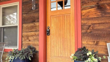 A wooden house with a red door, two large potted plants, a metal doorbell, and a concrete porch.