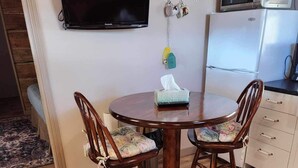 En matplats med ett runt träbord och två stolar, en tv som är monterad på väggen, en mikrovågsugn och ett kylskåp.