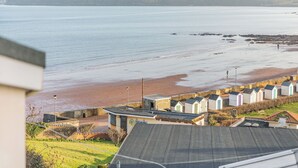 Vista costeira com uma praia, uma fileira de pequenas cabanas de praia e uma área gramada com um edifício em primeiro plano.