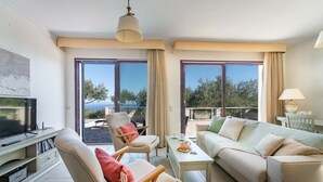 Un salon moderne avec un canapé, un fauteuil et une table basse. On y trouve un téléviseur posé sur un meuble, ainsi que de grandes fenêtres avec des rideaux.