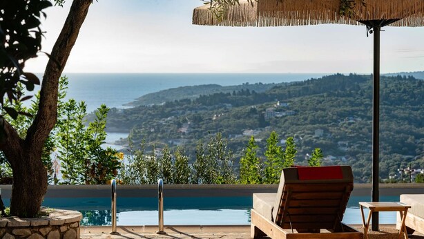 Un fauteuil de relaxation en bois avec un coussin rouge, une petite table en bois et une vue sur la piscine donnant sur un paysage côtier.