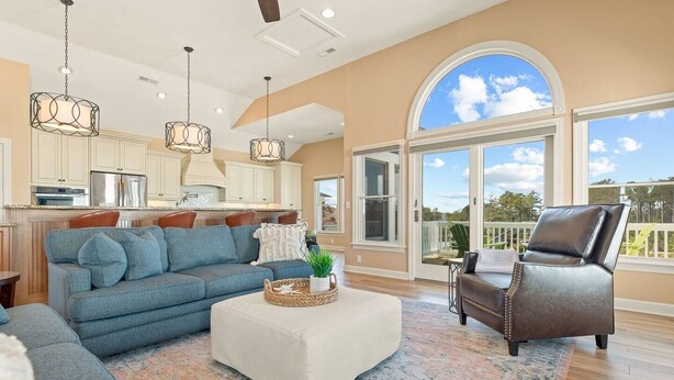 A spacious living room with a large sofa, a coffee table, and a leather chair. There is a kitchen area with white cabinets and a large window. - Waterfront,Pool,Game Room,Beach,DeepWaterAccess (Manteo)