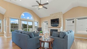 A spacious living room with two grey sofas, a wooden coffee table, a fireplace, and a large window overlooking a deck. - Waterfront,Pool,Game Room,Beach,DeepWaterAccess (Manteo)