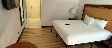 A hotel room with a bed, a desk, a chair, a TV mounted on the wall, and a door leading to another room.