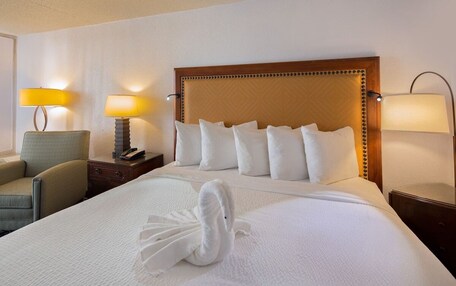 A neatly made bed with white linens and a swan-shaped towel, a wooden headboard, two bedside lamps, and a chair.