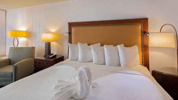 A neatly made bed with white linens and a swan-shaped towel, a wooden headboard, two bedside lamps, and a chair. - Home Inn Woods Hotel (Olive Branch)