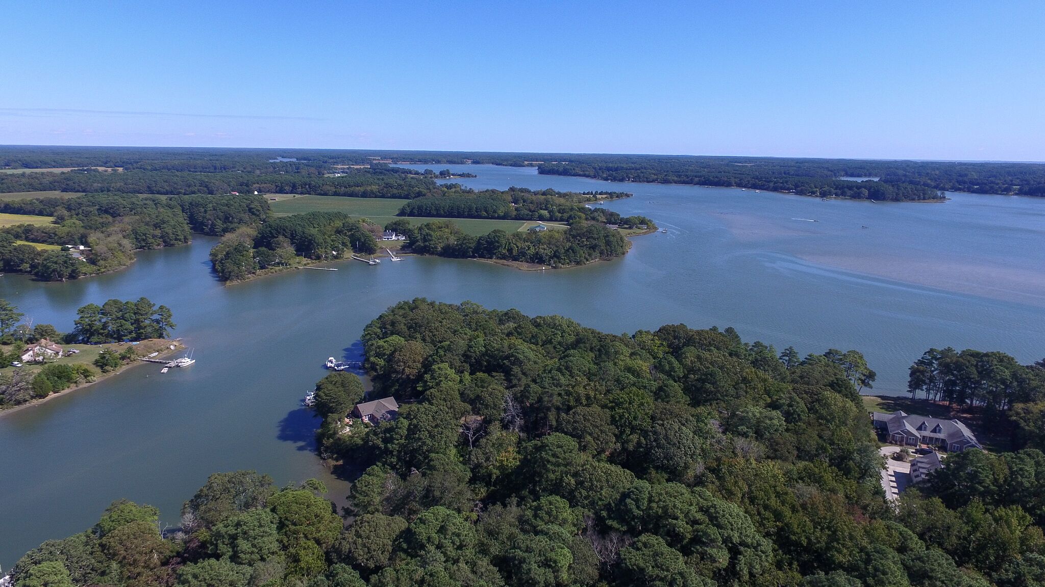 Vue aérienne d’une propriété au bord du lac, dotée de plusieurs quais, et entourée de forêts denses et de vastes champs.
