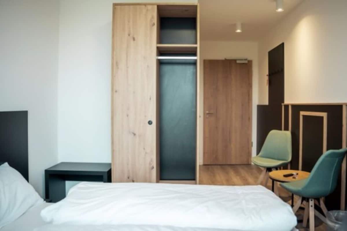 A modern hotel room with a bed, a wooden wardrobe, a small table with a chair, and a desk with a chair.