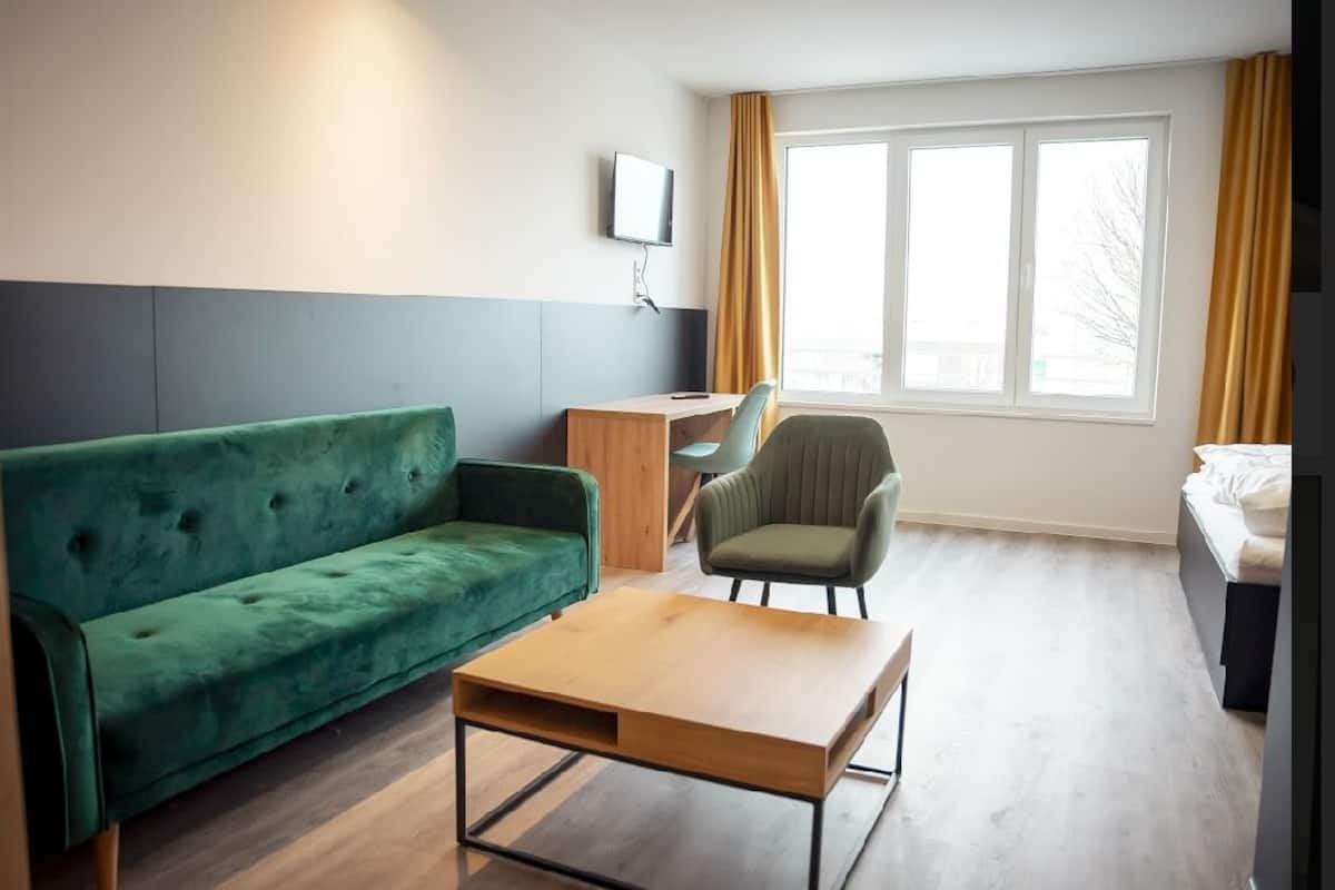 A modern hotel room with a green sofa, a wooden coffee table, a desk with a chair, a TV mounted on the wall, and a window with curtains.