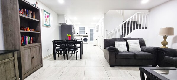 Una sala moderna con un sofá, una mesa de comedor con sillas, una estantería y una escalera que sube a otro nivel.