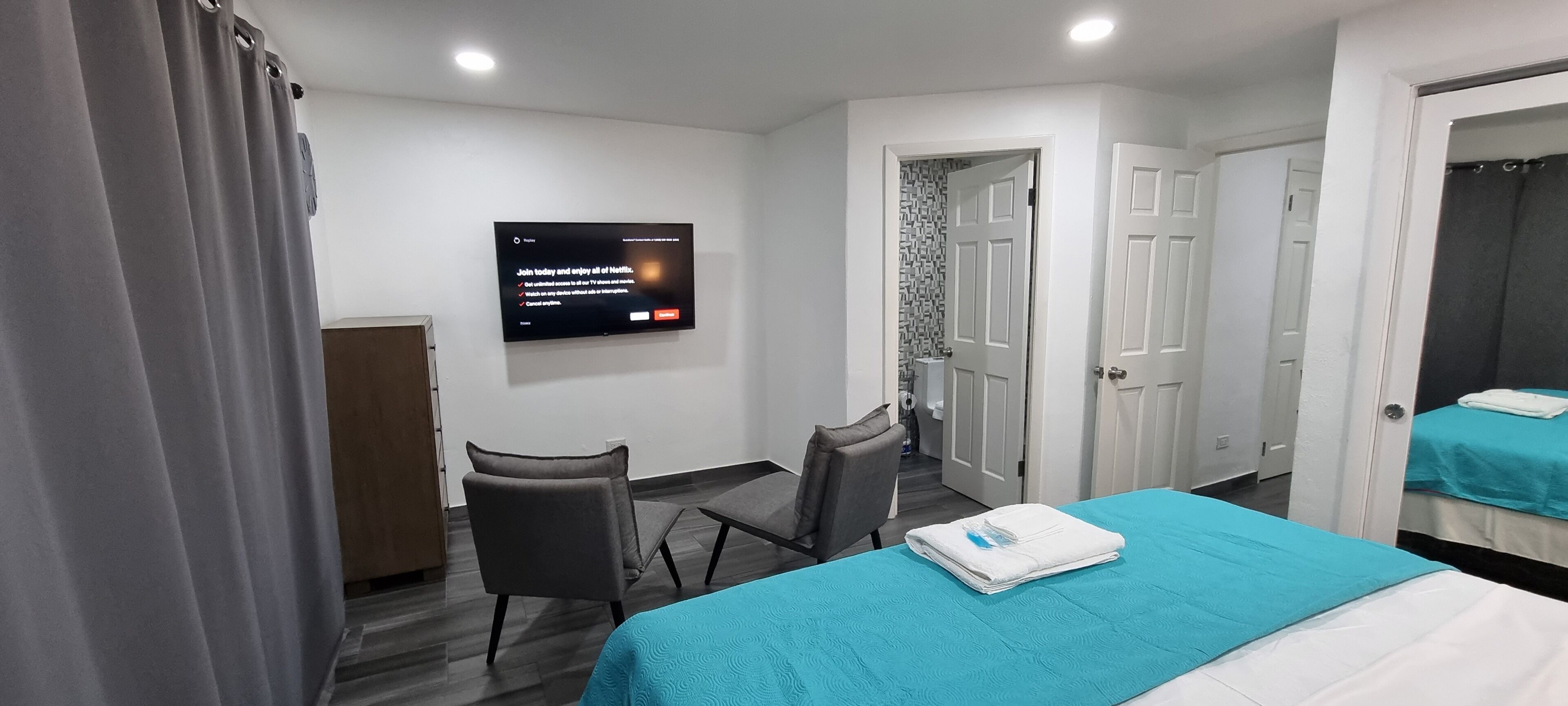 A hotel room with a bed, a desk, a chair, a TV mounted on the wall, and a bathroom visible through an open door.