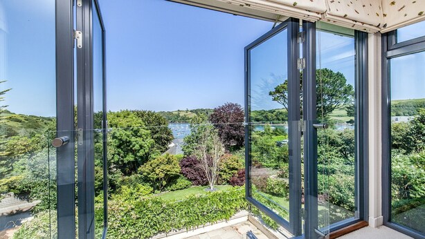 A balcony with large glass doors, offering a view of a lush garden and a body of water with boats.