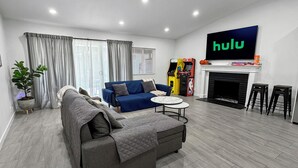 A modern living room with a grey sofa, a blue armchair, a coffee table, and a fireplace. A large TV screen displays the Hulu logo.