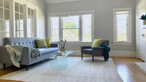 Un salon spacieux avec un canapé gris, un fauteuil assorti et une table basse en verre. De grandes fenêtres laissent entrer beaucoup de lumière naturelle.