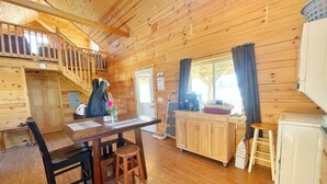 Interior de una cabaña de madera con comedor, cocina y una escalera que sube a un nivel superior.