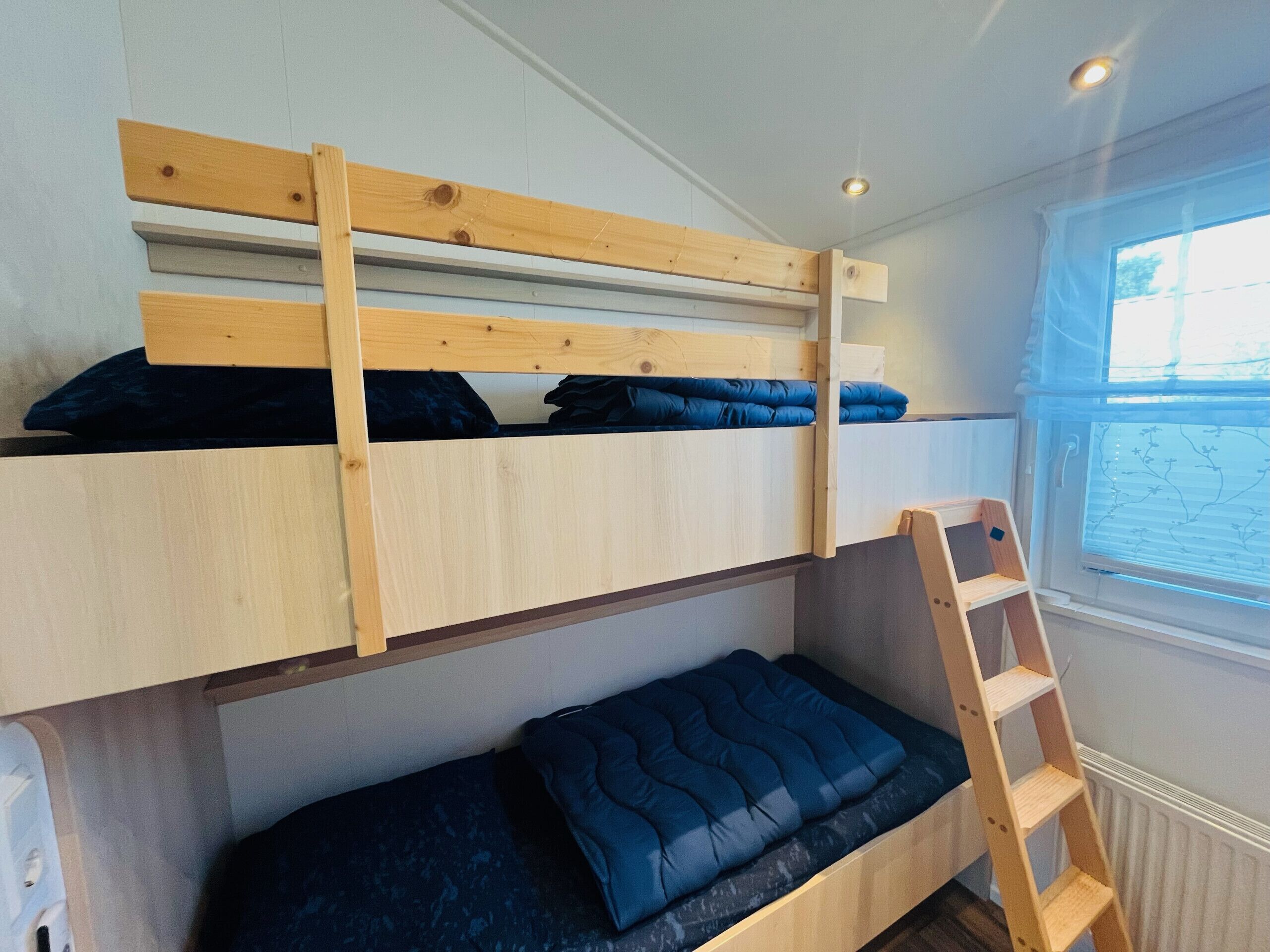 Ein Stockbett mit Holzleiter, ein Fenster mit Jalousien und eine Heizung.