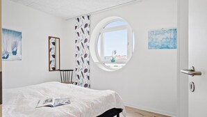 Een slaapkamer met een bed, een rond raam, een stoel en kunstwerken aan de muren.