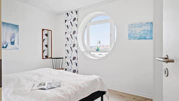 A bedroom with a bed, a round window, a chair, and artwork on the walls.