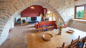 A rustic stone-walled room with a dining table, a TV, and a cozy seating area. - Gîte Aulas 2/4 Pers - LA Tieule (LA TIEULE)