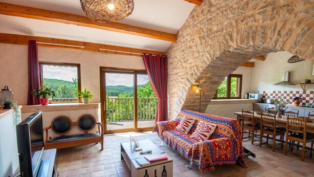 A cozy living room with a stone arch, a sofa, a TV, and a dining area with a kitchen in the background. - Gîte Aulas 2/4 Pers - LA Tieule (LA TIEULE)