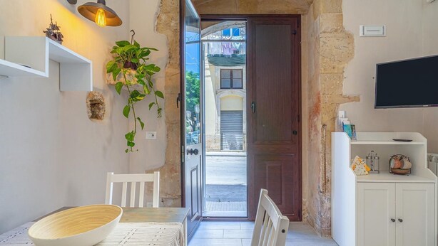 A dining area with a wooden table, a white chair, a hanging plant, and a white cabinet with a TV above it. - Lukìa House in the heart of Syracuse (Siracusa)