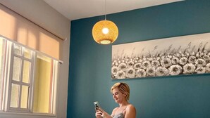 Una habitación con cama, ventana con persianas, lámpara colgante y un cuadro floral en la pared.