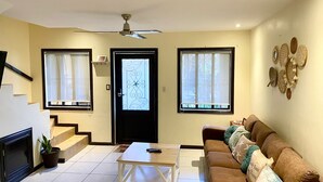 A living room with a sofa, coffee table, and a fireplace. There are two windows with curtains, a door with a decorative glass panel, and a ceiling fan.