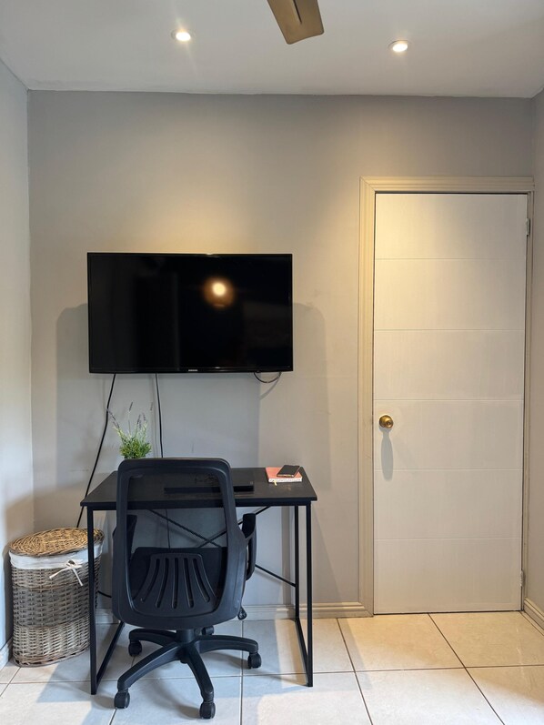 A room with a black office chair, a desk, a flat-screen TV mounted on the wall, a closed door, and a wicker basket on the floor.