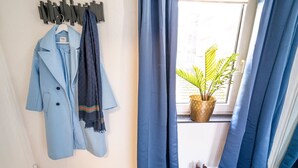 Une pièce comprenant un porte-manteau sur lequel est accroché un manteau bleu clair et une écharpe foncée, une plante en pot près d’une fenêtre et une paire de chaussures marron posées sur un tapis.
