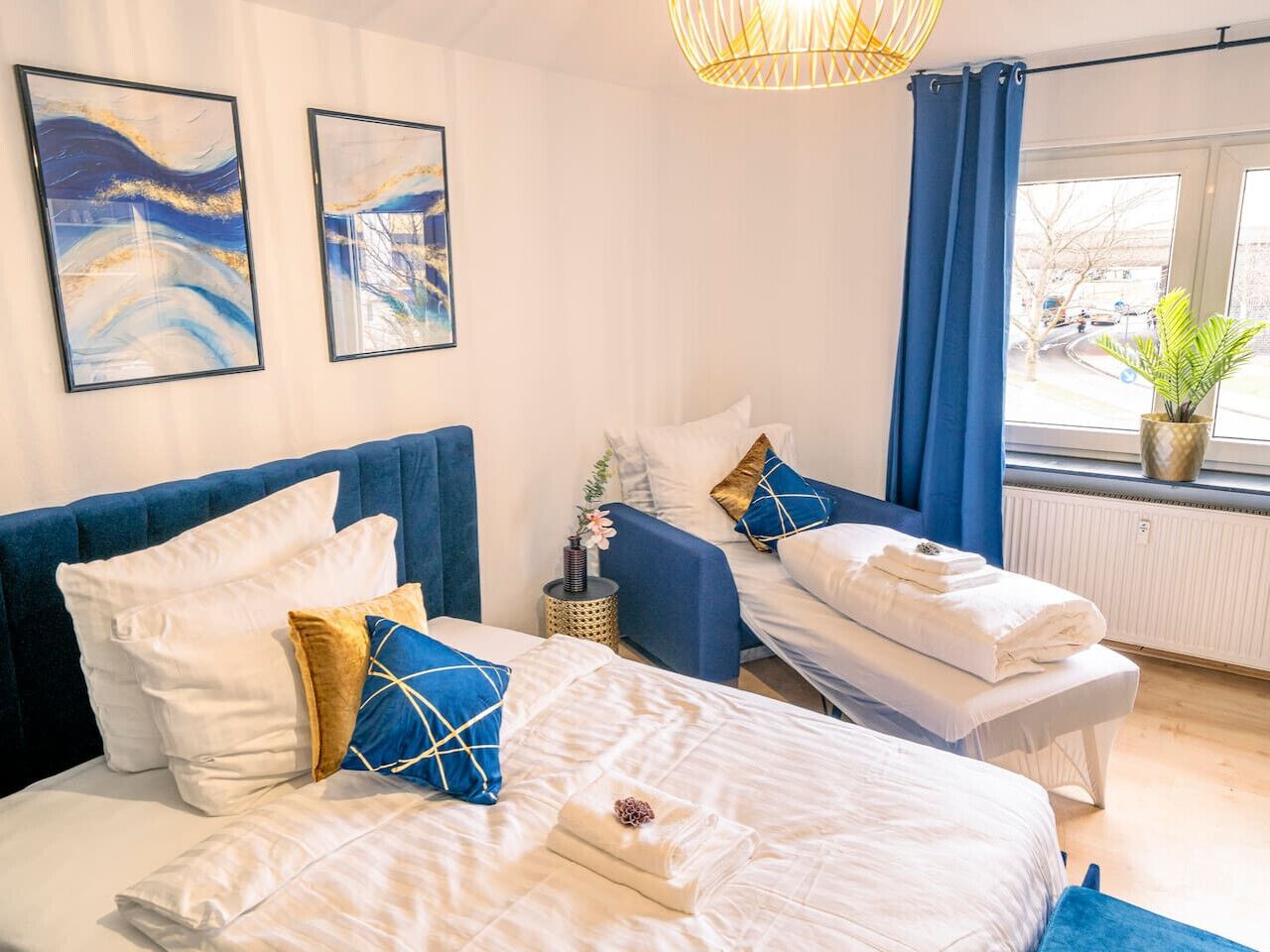 Une chambre d’hôtel avec deux lits, une fenêtre avec des rideaux bleus, une plante sur le rebord de la fenêtre et des œuvres d’art accrochées au mur.