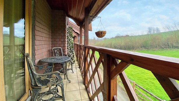 A wooden balcony with a table and chairs, a hanging basket, and a view of a green field. - 1 bedroom accommodation in Camerton, near Bath (Camerton, near Bath)