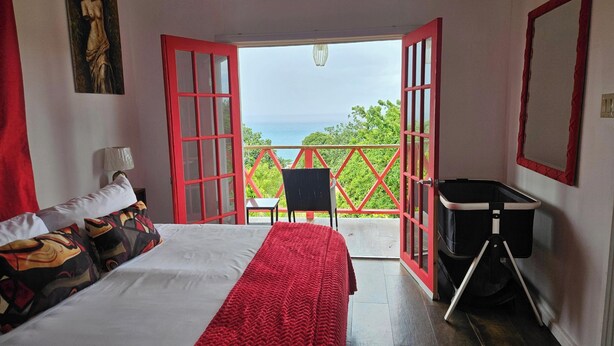Una stanza con un letto, una sedia, un tavolino, una finestra con cornice rossa e vista sul mare.