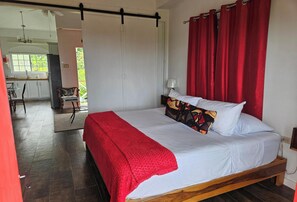 Ein Schlafzimmer mit Bett, roten Vorhängen, einem Holz-Kopfstück und Blick auf eine Küche und Essbereich.