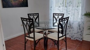 Zona pranzo con tavolo in vetro e quattro sedie, un quadro appeso alla parete, una finestra con tende leggere e pavimento piastrellato.