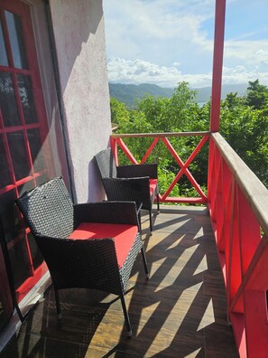 Un balcon avec deux fauteuils en osier, un coussin rouge et une vue sur des collines et des montagnes verdoyantes.