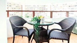 Een balkon met rieten stoelen en een tafel, een bloempot en uitzicht op hoogbouw.