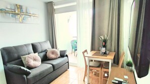 Een woonkamer met een donkergrijze bank, twee kussens, een houten eettafel met stoelen, een fles wijn en een vaas met bloemen.