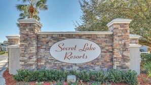 A sign for Secret Lake Resorts with a stone wall and colorful flower bed in front. - 🏰 10 min to Disney! Secret Lake Resort 3BDRM Condo 🌊Pool🔒Gated & Secure! (Kissimmee)