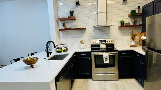 A modern kitchen with white subway tiles, black cabinets, stainless steel appliances, and wooden shelves. - 🏰 10 min to Disney! Secret Lake Resort 3BDRM Condo 🌊Pool🔒Gated & Secure! (Kissimmee)